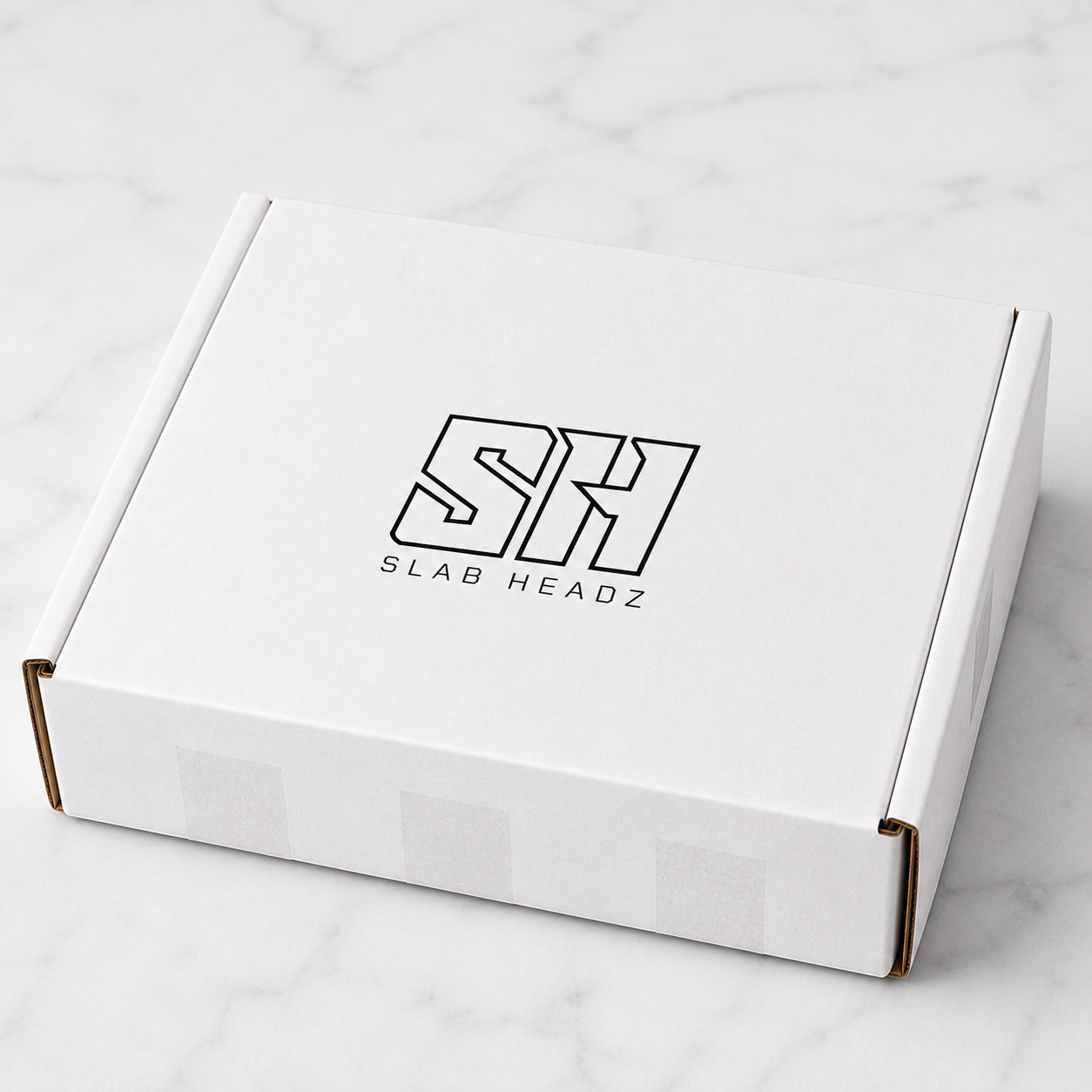 A white cardboard product box with a black outlined 'SH SLAB HEADZ' logo on the lid, photographed on a light marble surface.